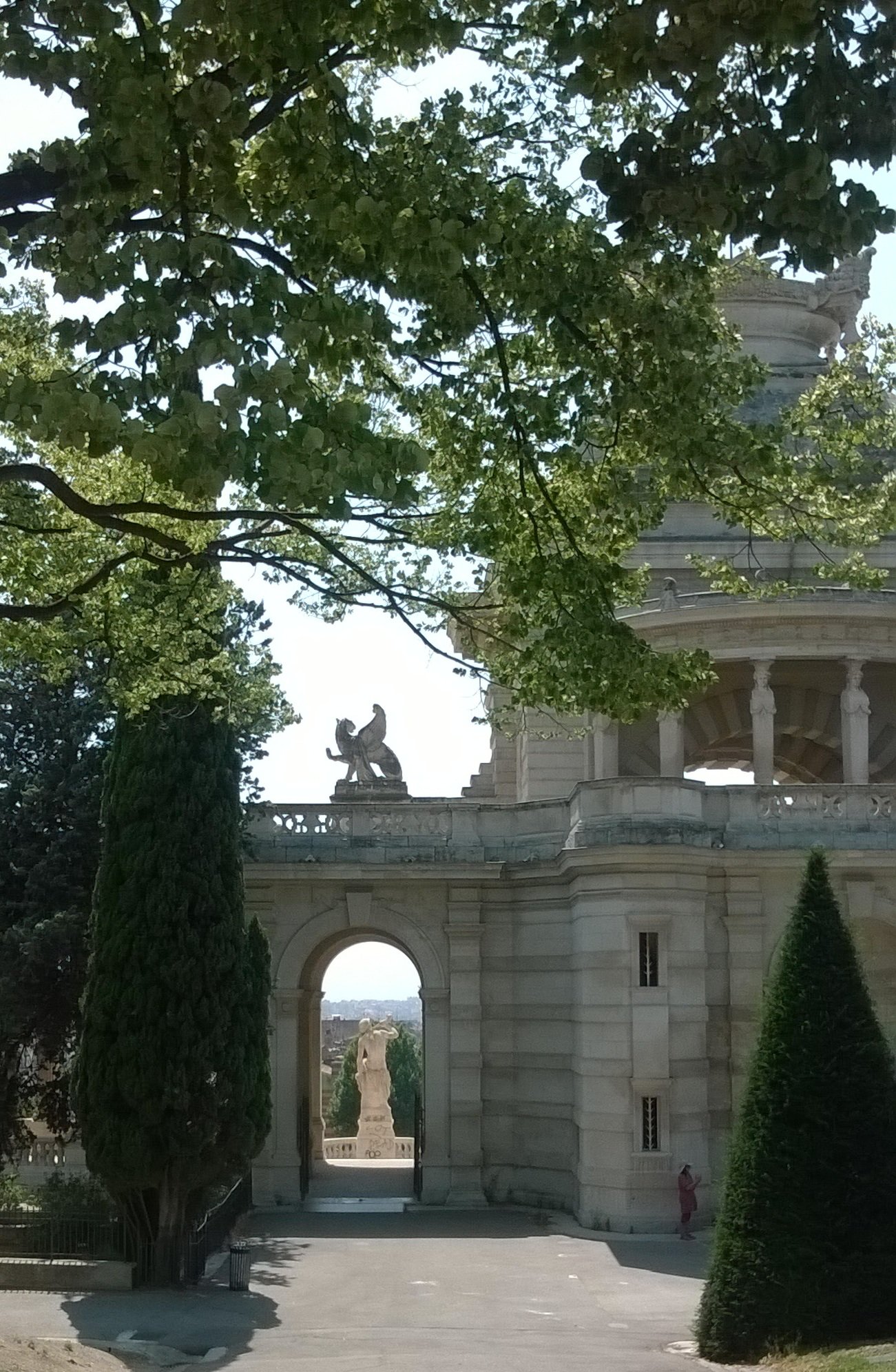 au premier plan des branche d'arbre plus loint l'arrière du monument du parc Longchamp avec ses colonnes et son phénix, tout au fond la statue de la Durance et la ville